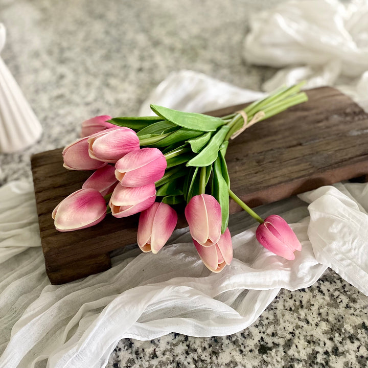 A bundle of mauve real touch tulips tied with a paper bow, placed on a wooden surface with a white translucent fabric beside it.