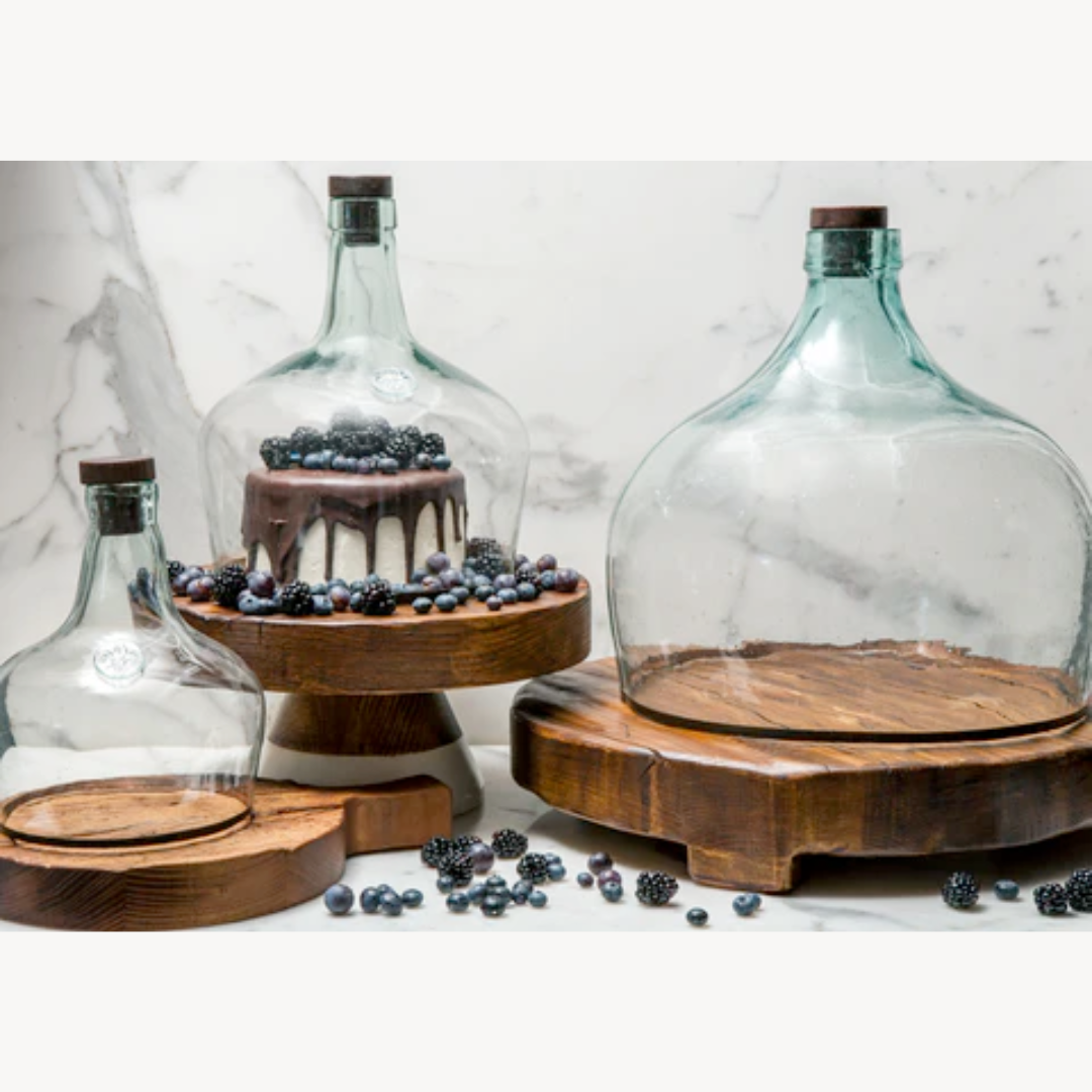 Glass cloches with wooden bases on a marble surface, containing berries and a small cake.