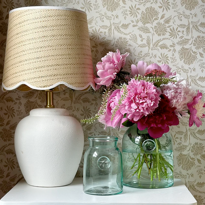 White lamp with beige shade next to pink flowers in a clear French mason vase on a floral-patterned wall.