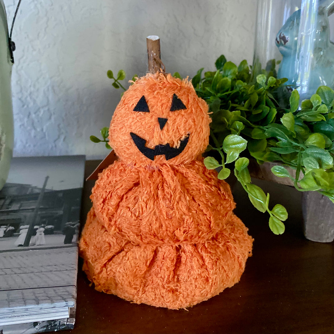 A stack of three orange fleece pumpkins with black felt shapes for the face, a wooden stick, and dried vines on top, designed as a Halloween decoration.