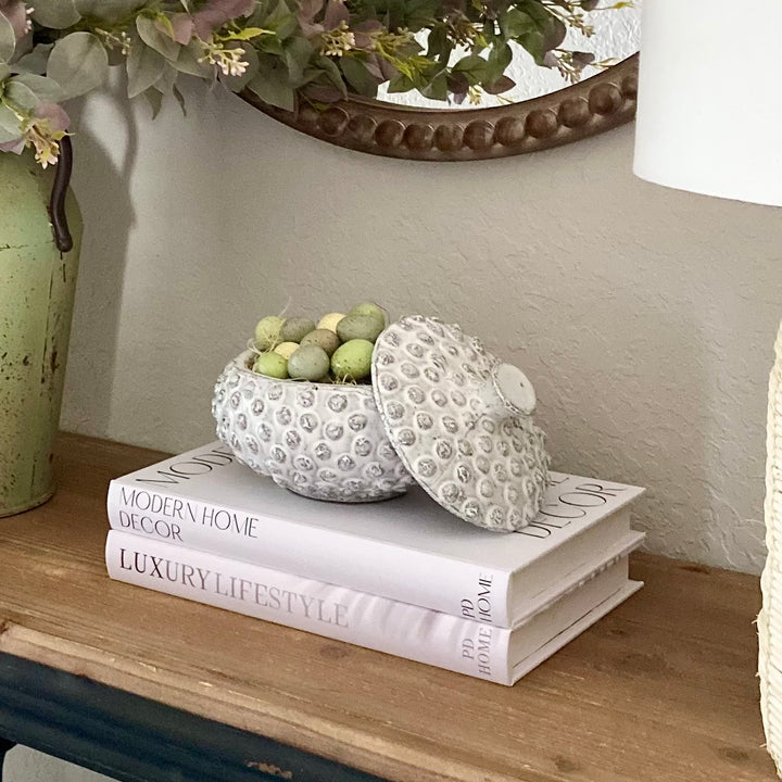 A set of three decorative storage books with faux book covers, placed on a table with decorative items on top.