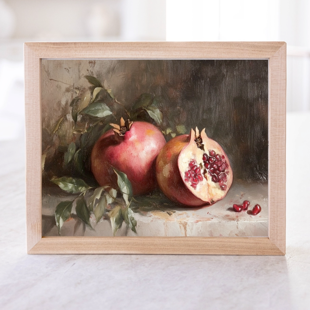 A framed artwork depicting a pomegranate cut in half with seeds scattered around, set against a dark background.