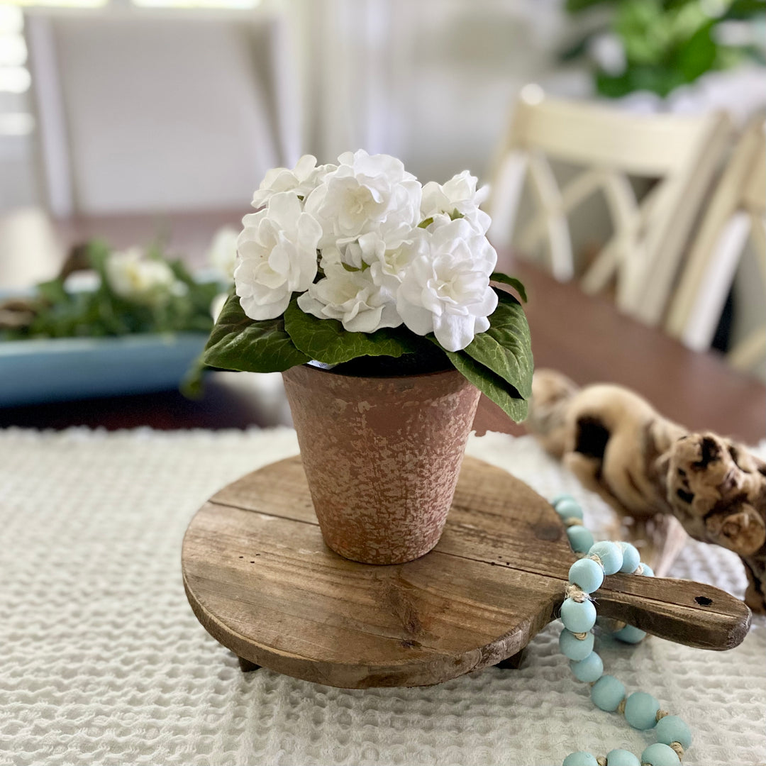 An artificial White Violet in a Distressed Clay Pot, placed on a wooden round tray with a decorative blue and white object to the side.