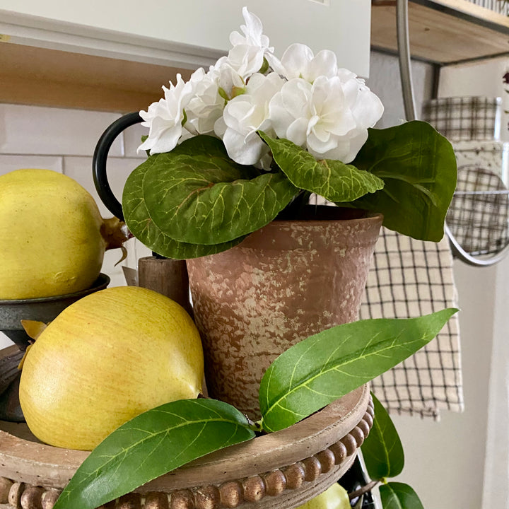 An artificial White Violet plant with green leaves in a distressed clay pot, placed on a decorative tray with faux pomegranates.
