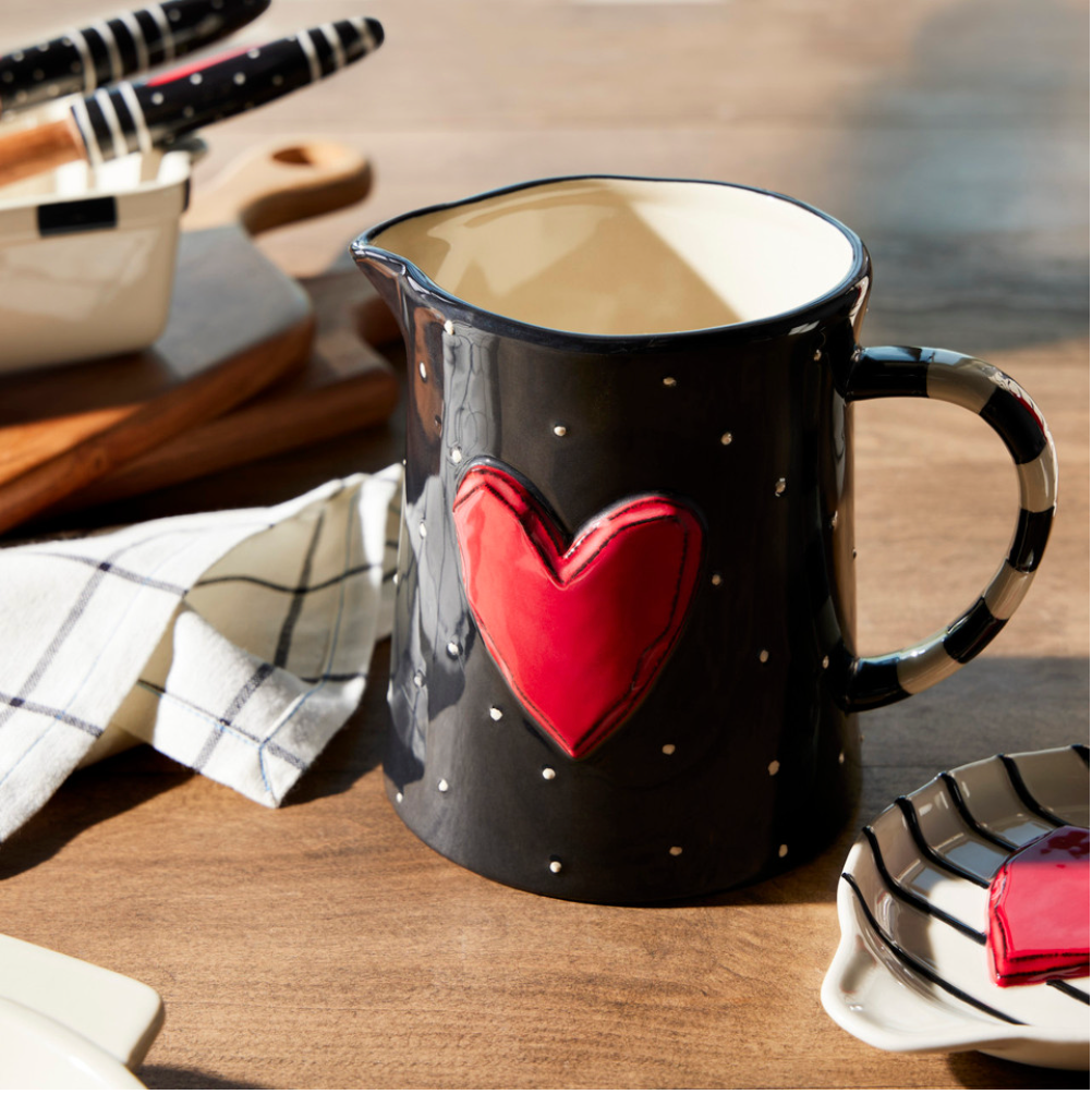 A black and white pitcher with a red heart design and white polka dots on a table with a checkered napkin and wooden cutlery in the background.
