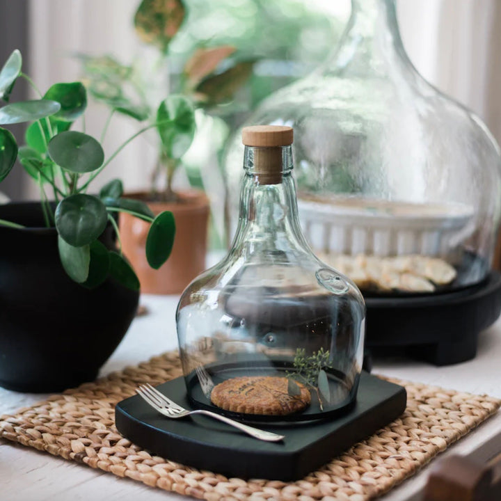 A clear glass demijohn cloche with a black distressed rim and wood cork, displayed on a woven mat with a plant in the background.