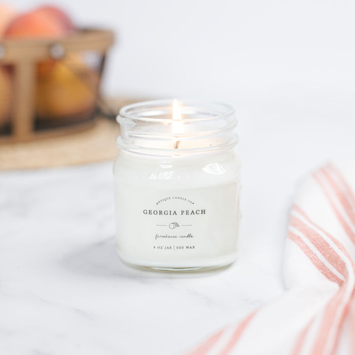 A Georgia Peach scented candle in a clear glass jar with a metal lid, placed on a white surface with a striped red and white cloth to the side and a basket of fruit in the background.
