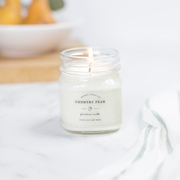 A lit Country Pear scented candle in a clear glass mason jar with a metal lid, placed on a white surface with a bundle of dried oranges in the background.