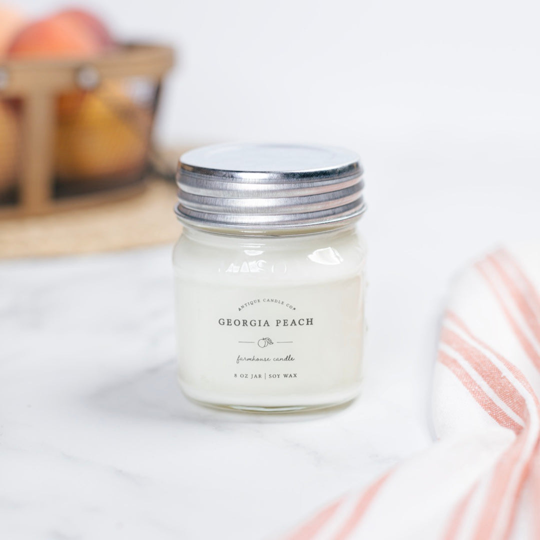 A Georgia Peach scented candle in a clear mason jar with a metal lid, placed on a table with a striped napkin and fruits in the background.