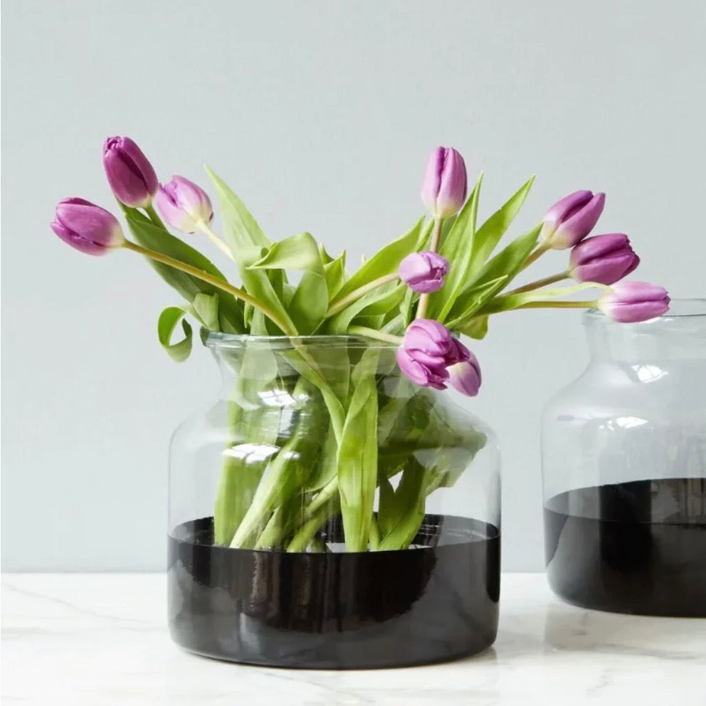Black color block glass vase holding orange tulips with green stems, set on a white marble surface against a white background.