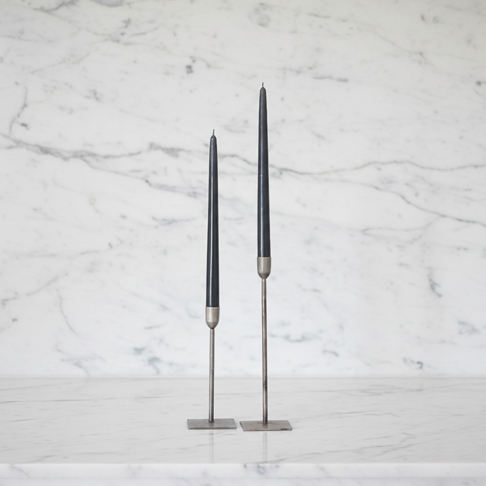 Two slim charcoal gray taper candles in silver metal holders, standing on a white marble surface against a white marble background.