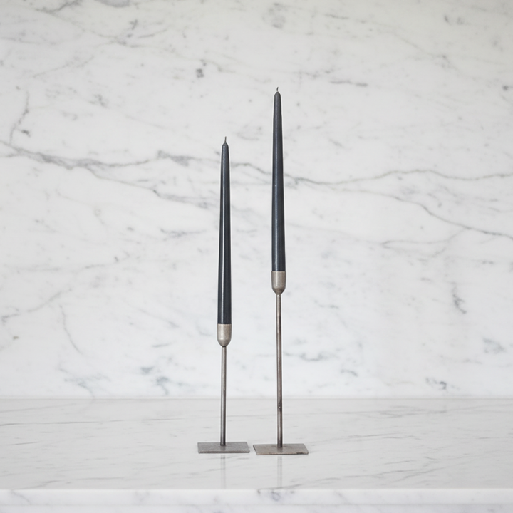 Two slim charcoal gray taper candles in silver metal holders, standing on a white marble surface against a white marble background.