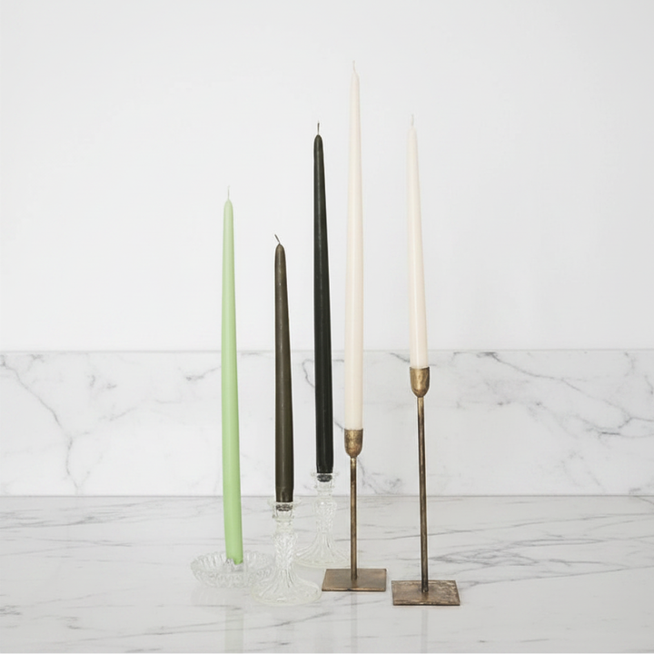 A selection of 12-inch taper candles in ivory and gray, displayed in various candle holders against a neutral background.