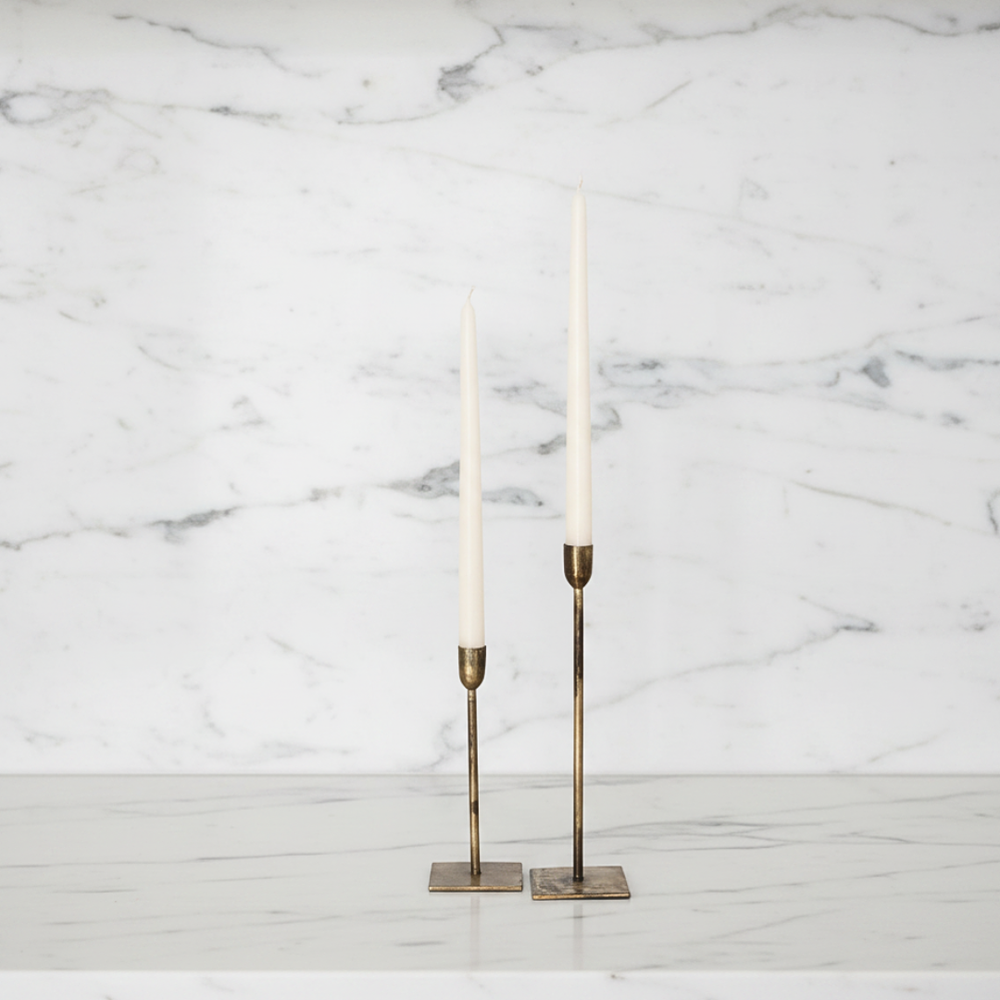Two slim brass candlesticks holding ivory taper candles, displayed on a white marble surface against a white marble wall background.