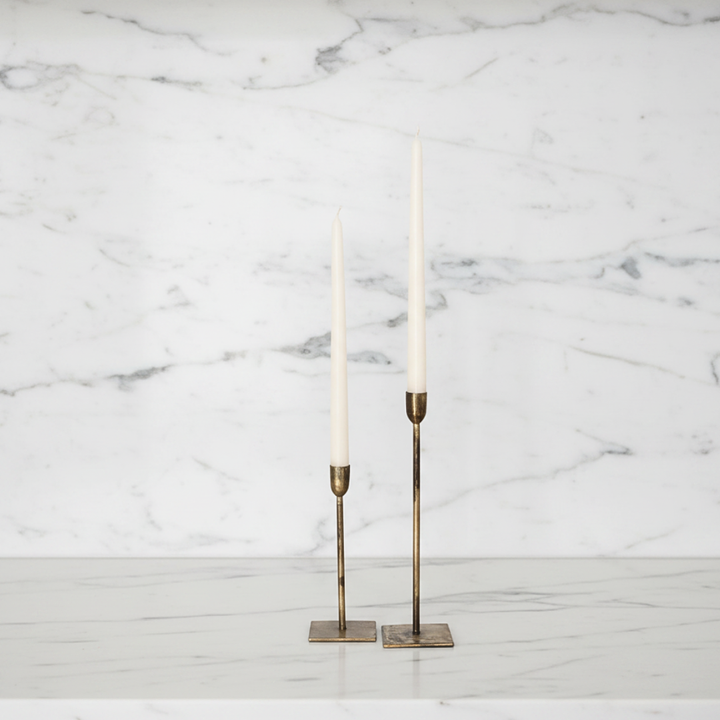 Two slim brass candlesticks holding ivory taper candles, displayed on a white marble surface against a white marble wall background.