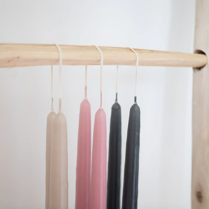 A selection of 12-inch taper candles in beige, pink, black, and gray, displayed hanging on a wooden rod against a neutral background.