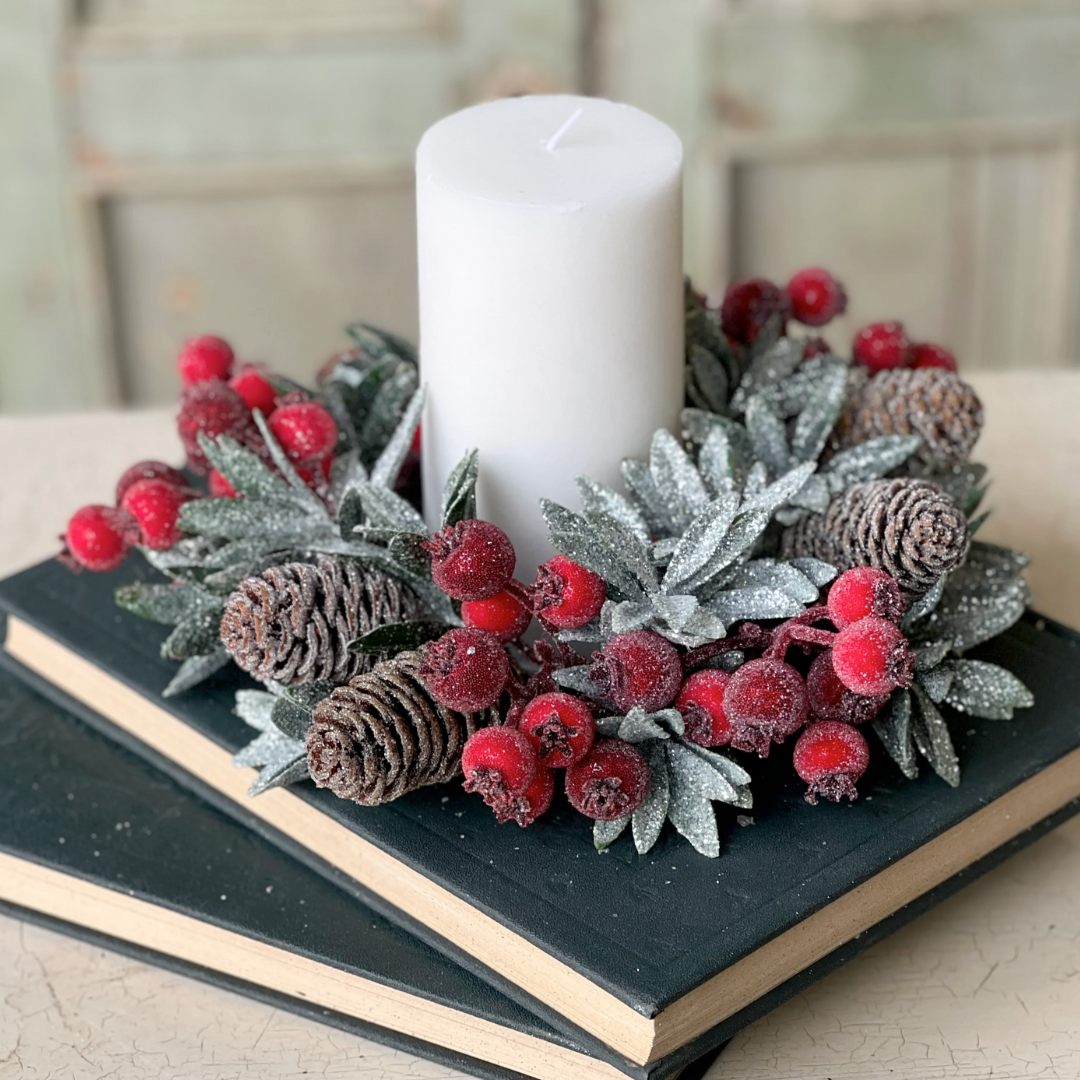 A decorative candle ring featuring sugary laurel leaves, glitter, and icy pine cones, with a central candle, surrounded by red berries on a black book-like surface.