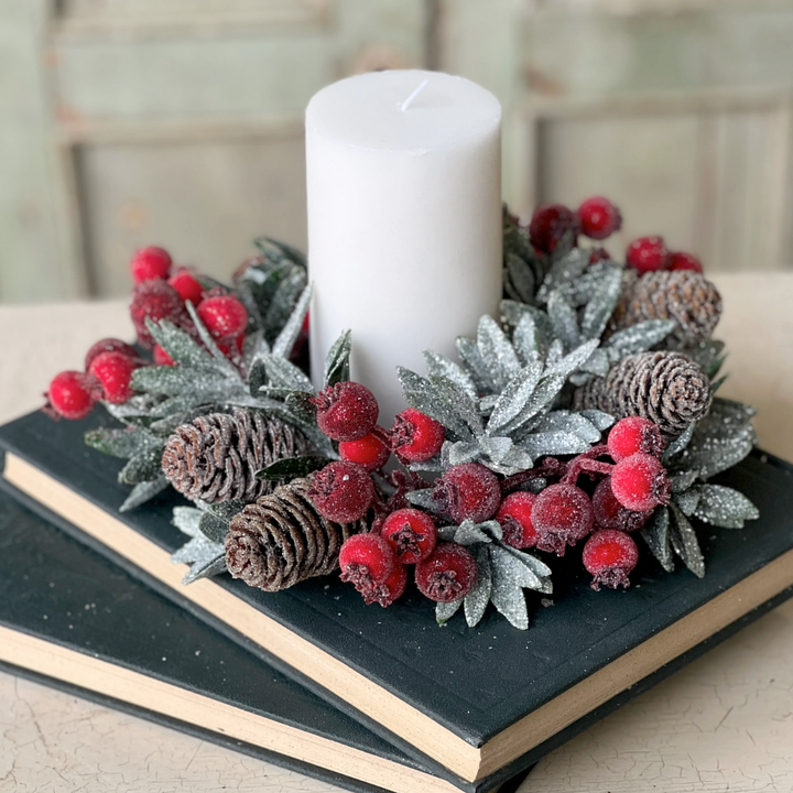 A decorative candle ring featuring sugary laurel leaves, glitter, and icy pine cones, with a central candle, surrounded by red berries on a black book-like surface.