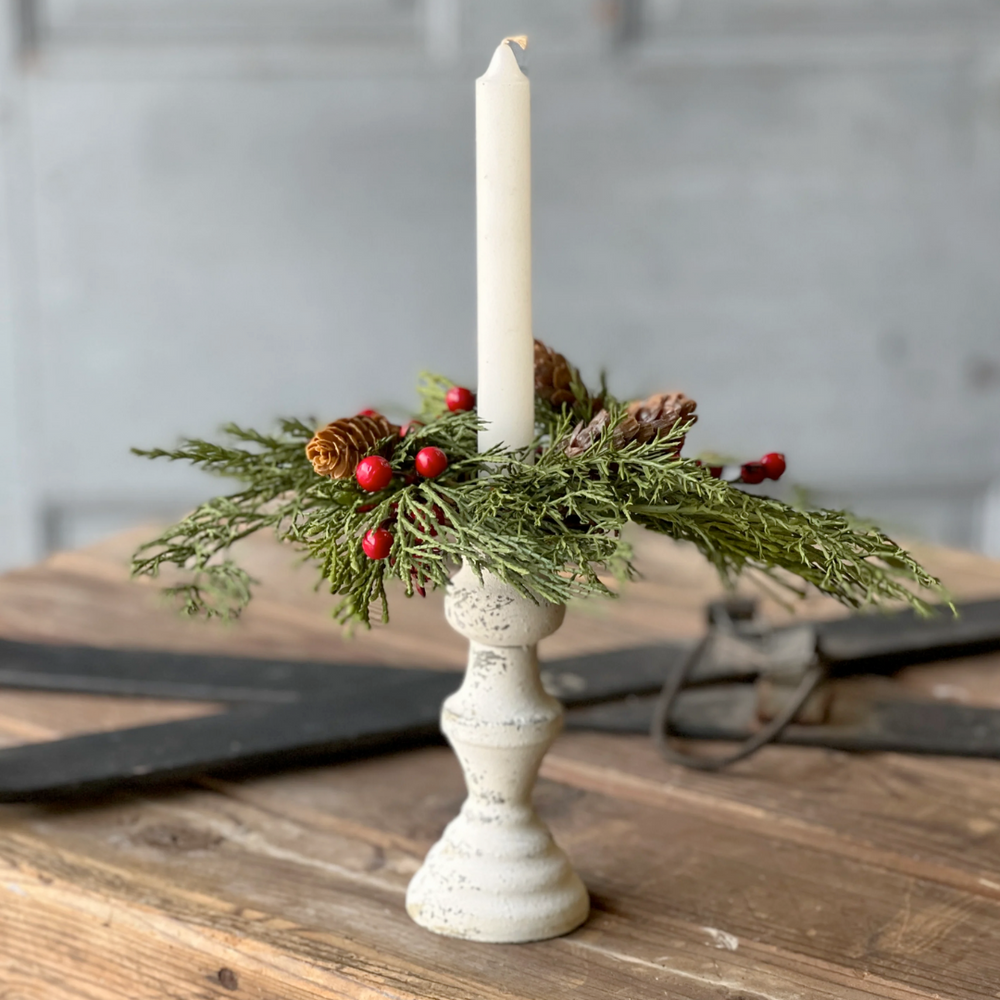 A decorative Alpine with berries candle ring made of plastic, styrofoam, and pine cones, featuring a white grapevine base, displayed with a lit candle.