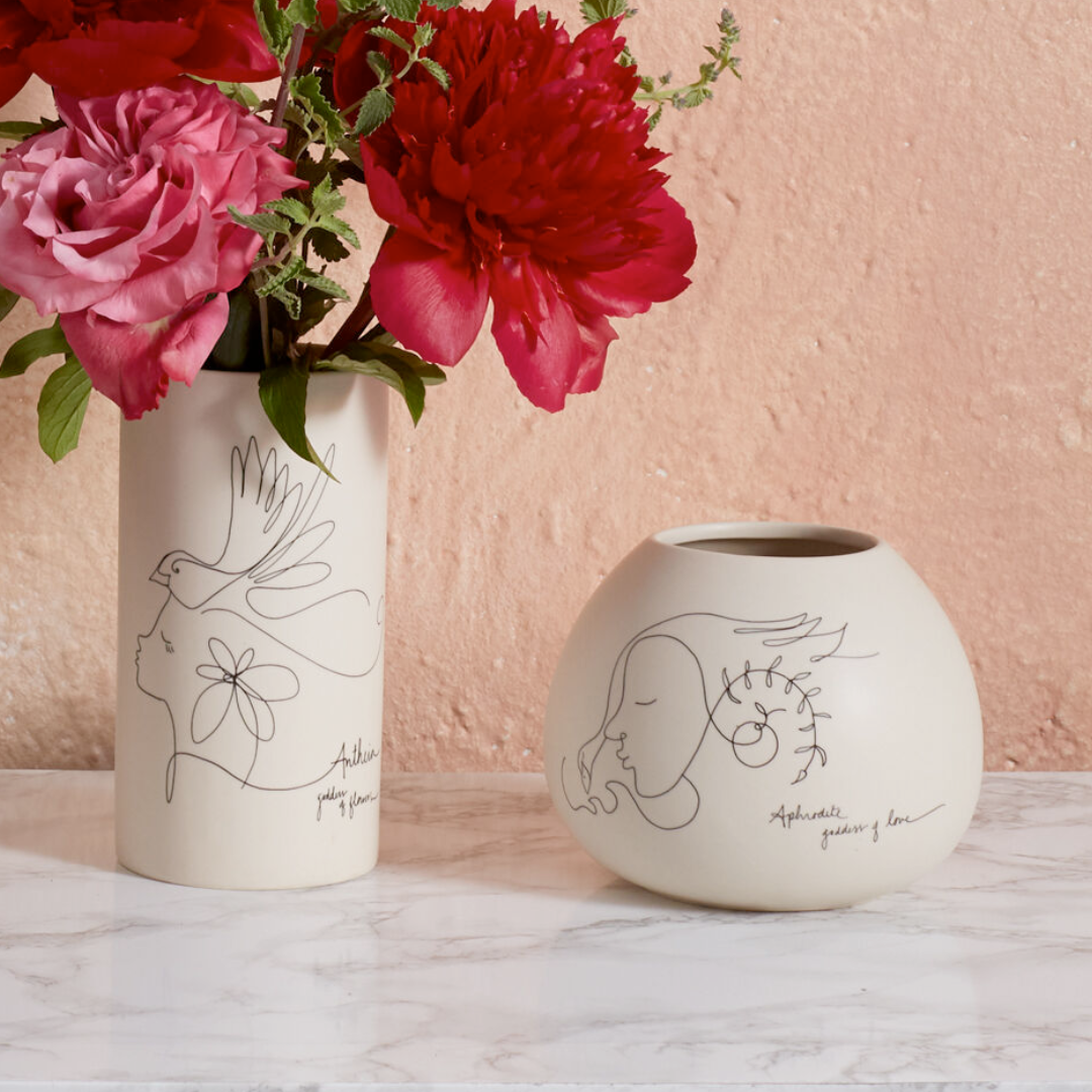 Two ceramic vases with line drawings of a goddess, one tall and slender and the other short and bulbous, with flowers arranged in the taller vase, displayed on a marble surface against a pink wall.