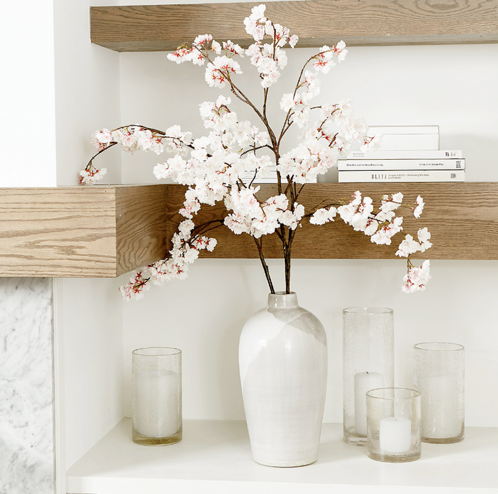 White vase with cherry blossom branches on a wooden shelf with candles and books.