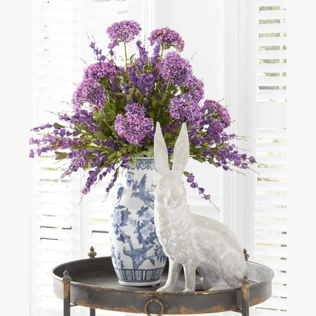 A decorative artificial Deep Purple Lavender Spray with lush green foliage in a blue and white vase, displayed on a metal tray with a white rabbit figurine.