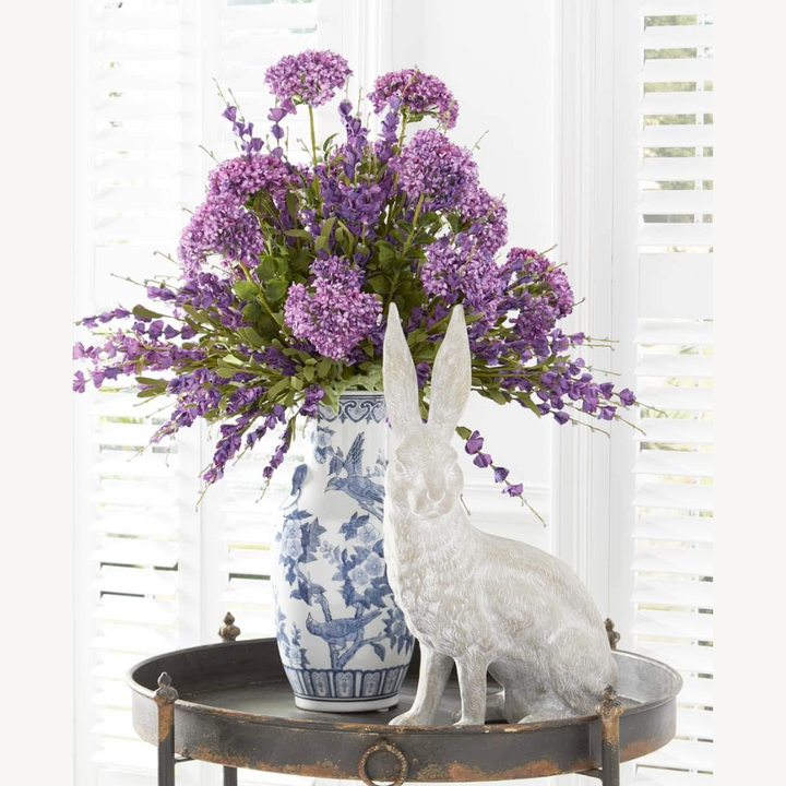 A decorative artificial Deep Purple Lavender Spray with lush green foliage in a blue and white vase, displayed on a metal tray with a white rabbit figurine.