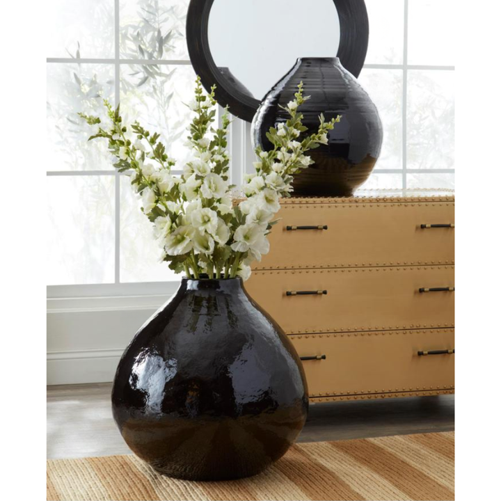 Black vase with white flowers on a wooden floor with a dresser in the background