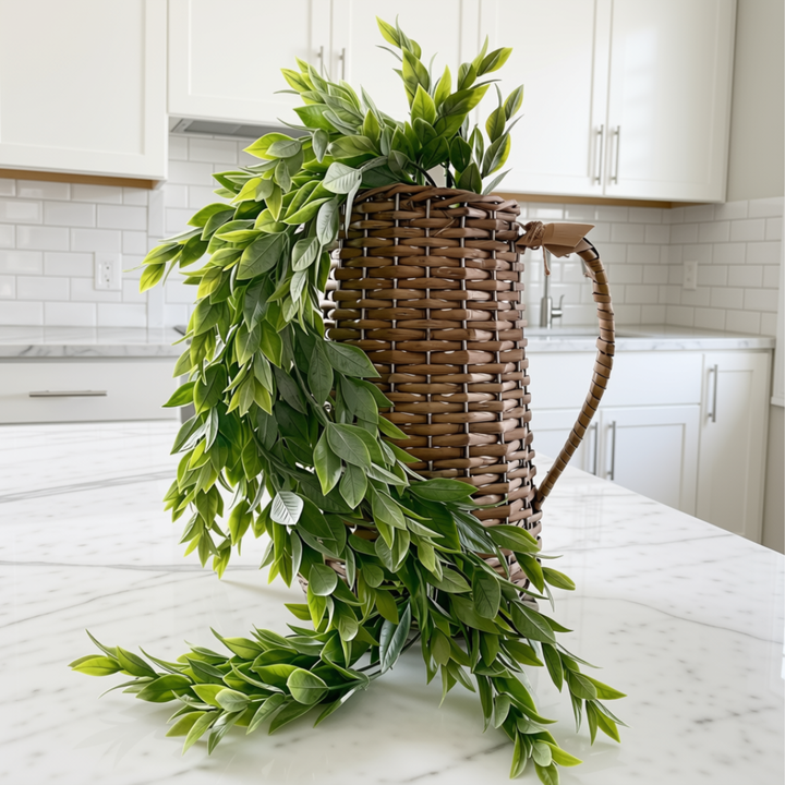 A decorative artificial green leaf garland draped over a brown woven basket with a handle, placed on a white marble kitchen counter.