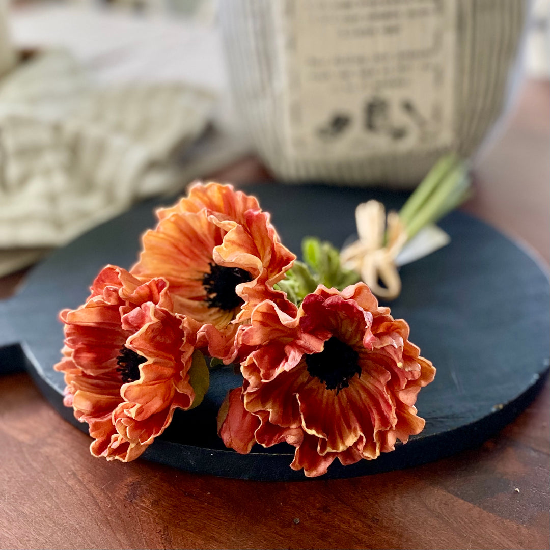 A bundle of artificial poppy flowers in shades of orange and red with raffia tie, displayed on a dark surface.