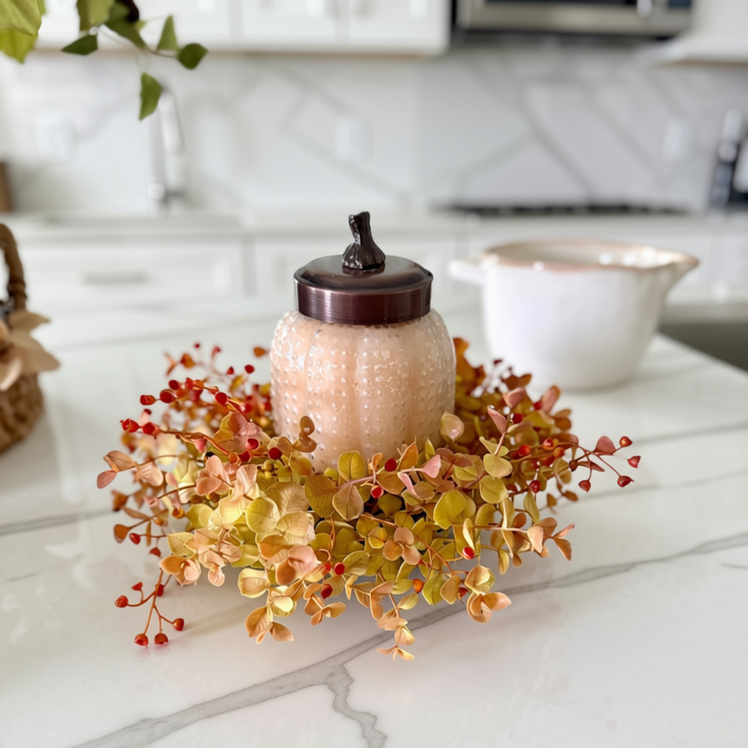 A decorative glass jar with a dark brown lid sits on a white marble countertop, surrounded by an autumn floral arrangement with yellow, orange, and red leaves.