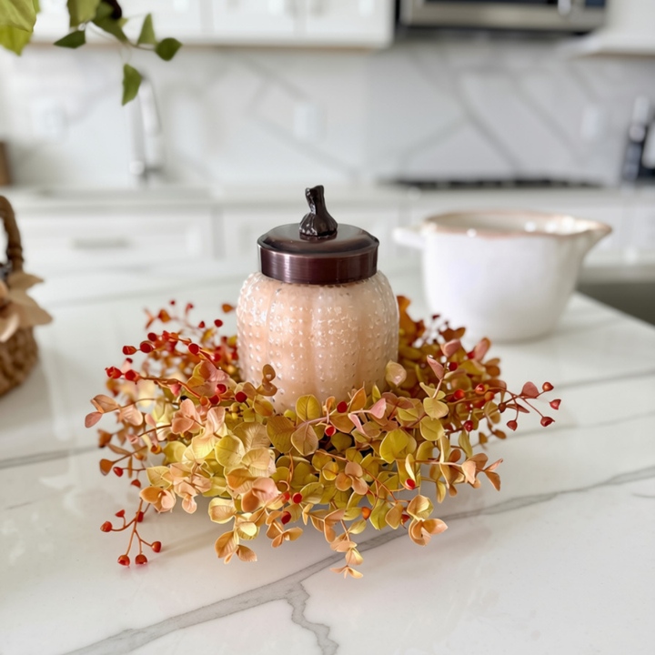 A decorative glass jar with a dark brown lid sits on a white marble countertop, surrounded by an autumn floral arrangement with yellow, orange, and red leaves.