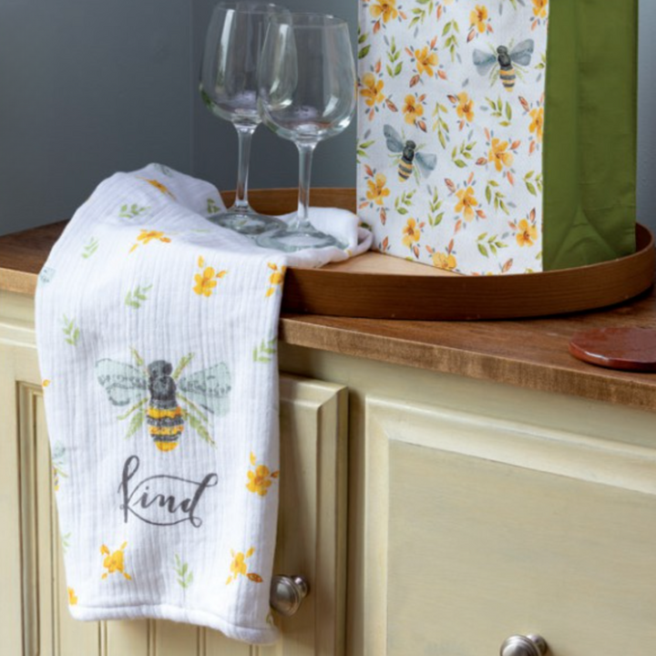 A white hand towel with a bee and flower design, labeled 'KIND', hung on a wooden rack next to two glasses and a green bag.