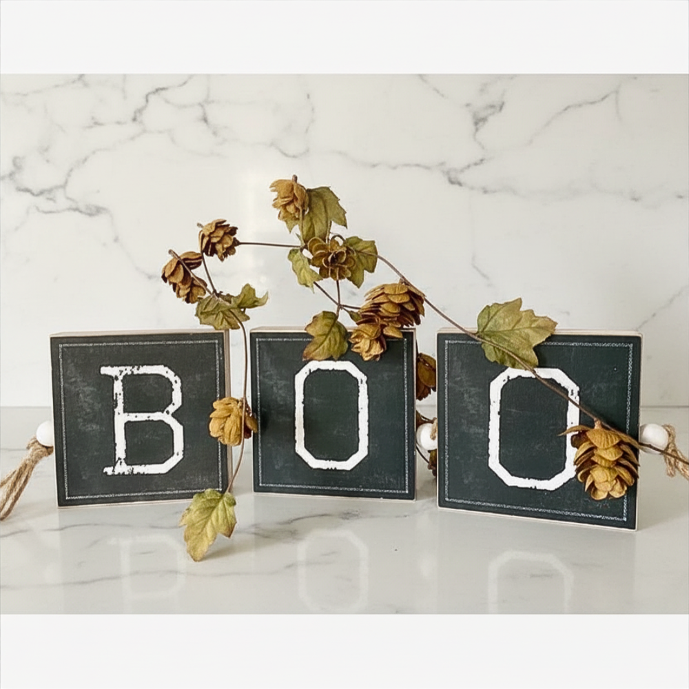 Decorative sign with 'BOO' letters surrounded by autumn leaves on a wooden surface.