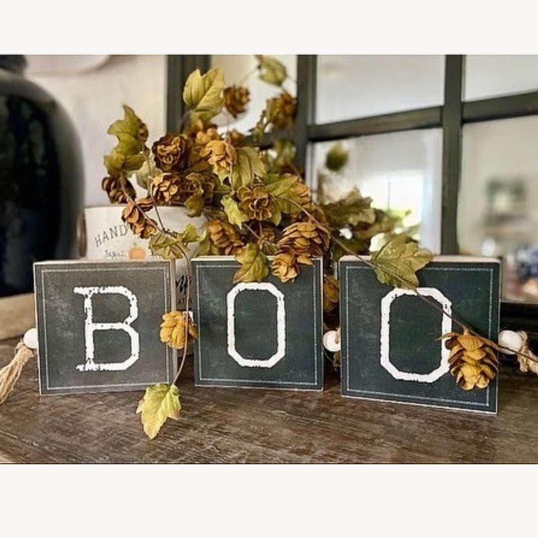Decorative sign with 'BOO' letters surrounded by autumn leaves on a wooden surface.