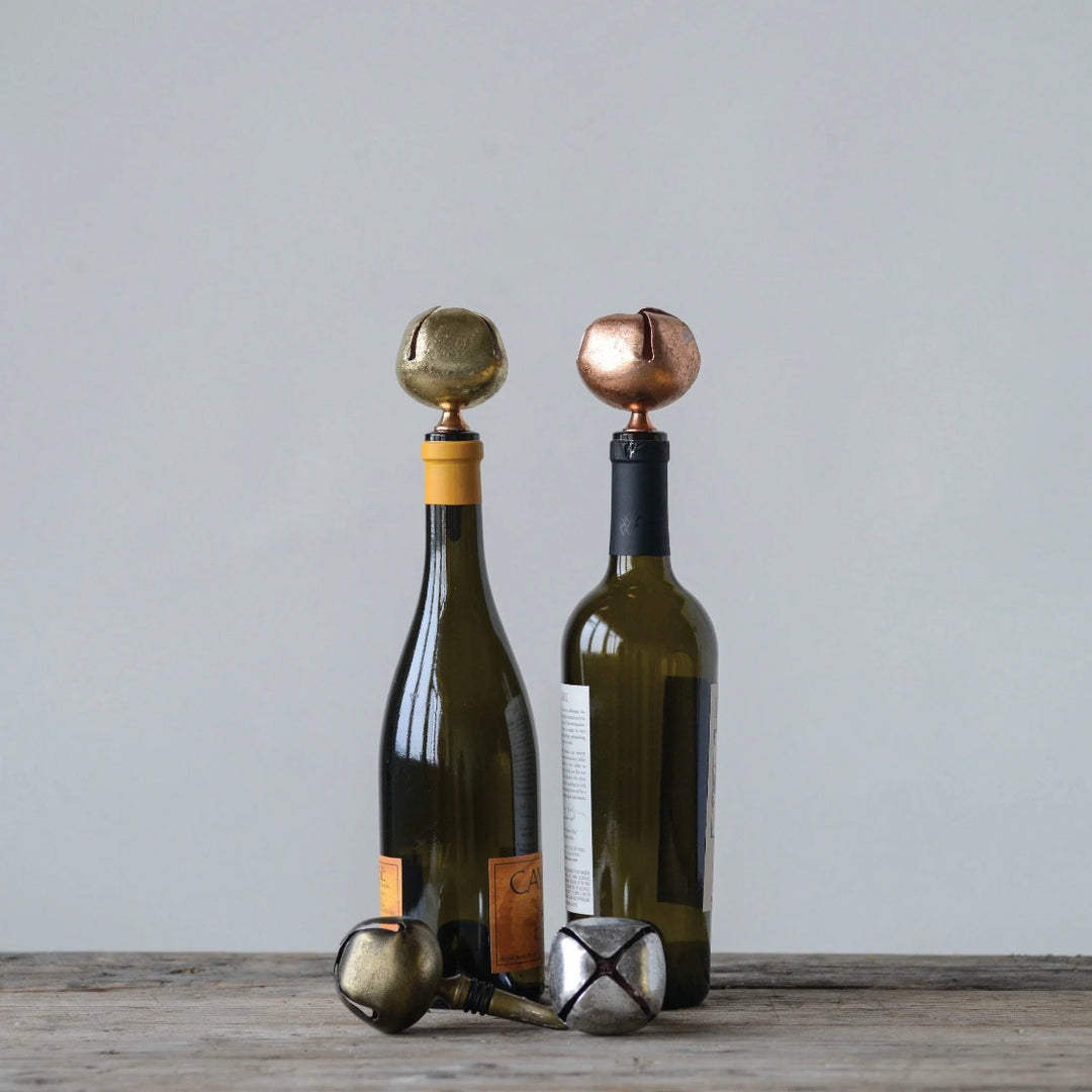 Two wine bottle stoppers with jingle bells, one in silver and one in gold, placed on a wooden surface with a wine bottle in the background.