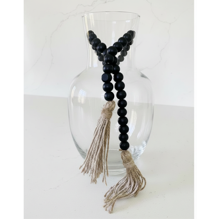 Black beaded garland with tassels hanging from a clear glass vase on a white background