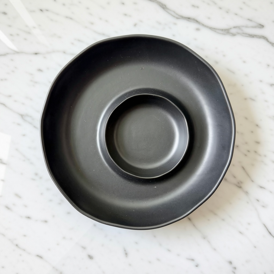 A matte black ceramic chip and dip serving plate with a scalloped rim, photographed from directly above on a white marble surface.