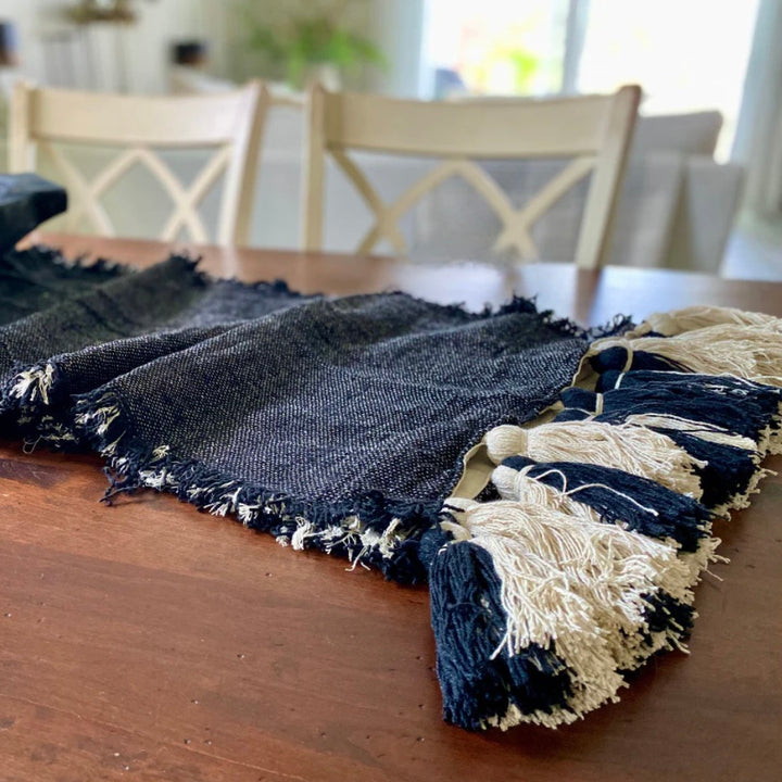 A black and ivory table runner with fringe trim and large chunky tassels is displayed on a wooden table.