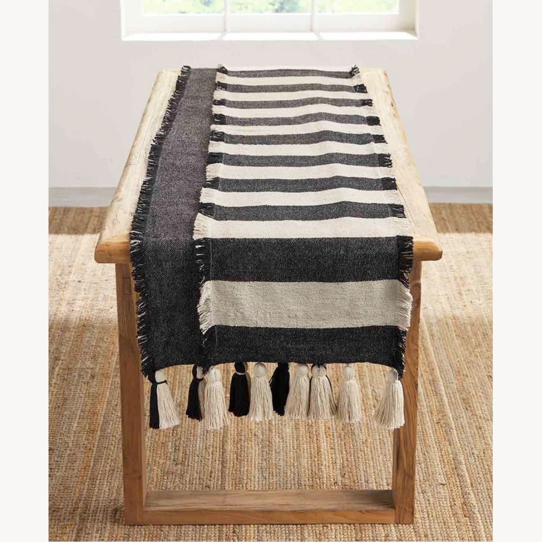 A black and ivory table runner with fringe trim and large chunky tassels is displayed on a wooden table.