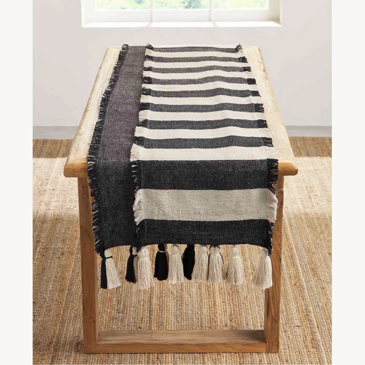 A black and ivory table runner with fringe trim and large chunky tassels is displayed on a wooden table.
