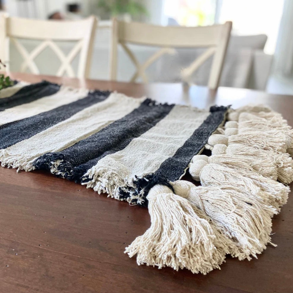 A black and ivory table runner with fringe trim and large chunky tassels is displayed on a wooden table.