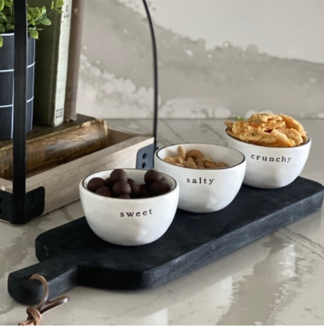 Three white bowls labeled 'sweet', 'salty', and 'crunchy' on a black tray with snacks.