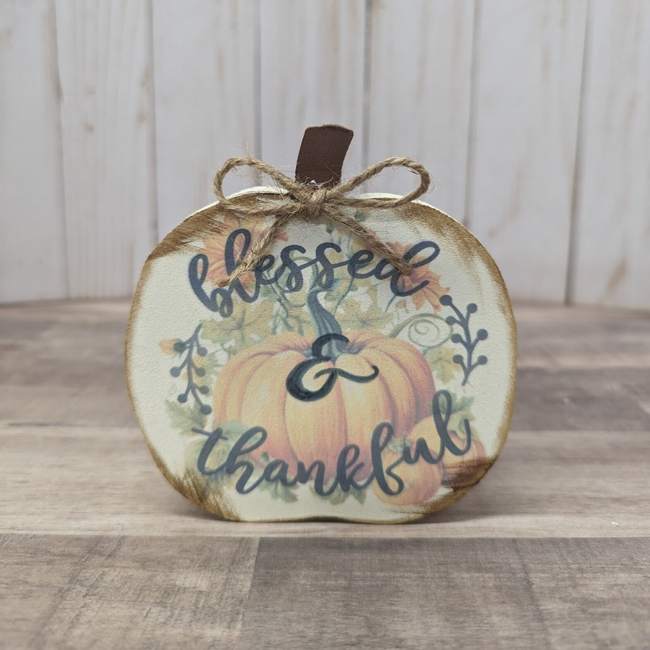 A round autumn-themed decorative plaque with a painted pumpkin, the words 'blessed & thankful' in dark blue script, and a small brown hanging loop tied with twine, set on a wooden surface against a light wood plank background.