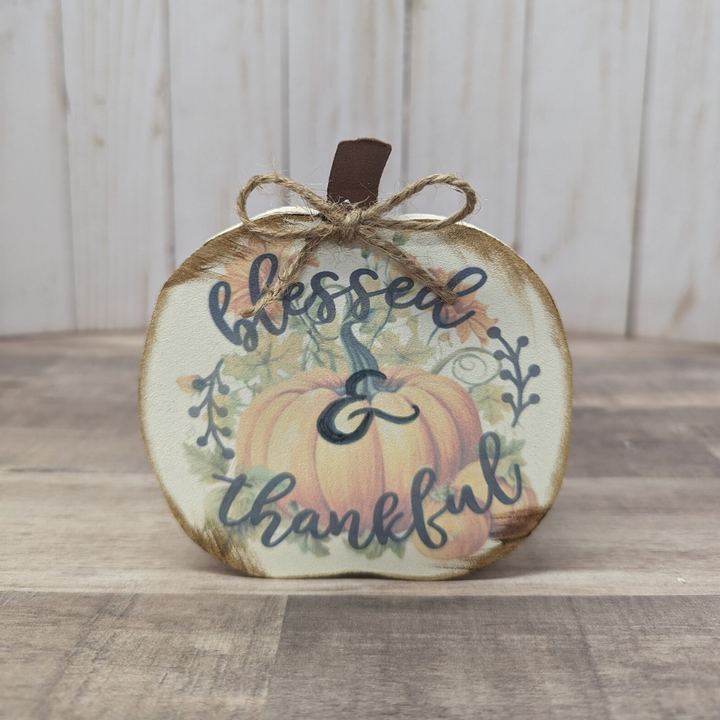 A round autumn-themed decorative plaque with a painted pumpkin, the words 'blessed & thankful' in dark blue script, and a small brown hanging loop tied with twine, set on a wooden surface against a light wood plank background.
