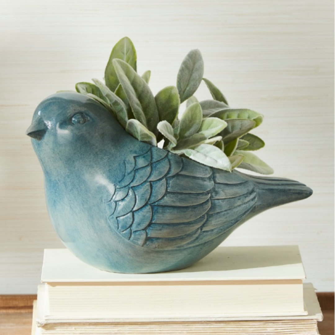 A blue bird-shaped planter made of resin, containing artificial green foliage, placed on top of a stack of books.
