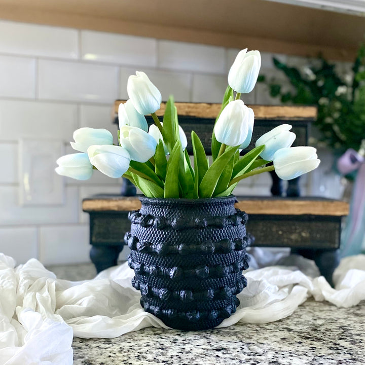 A bundle of artificial French Blue Real Touch Tulips arranged in a black textured knot cement vase, placed on a kitchen countertop.