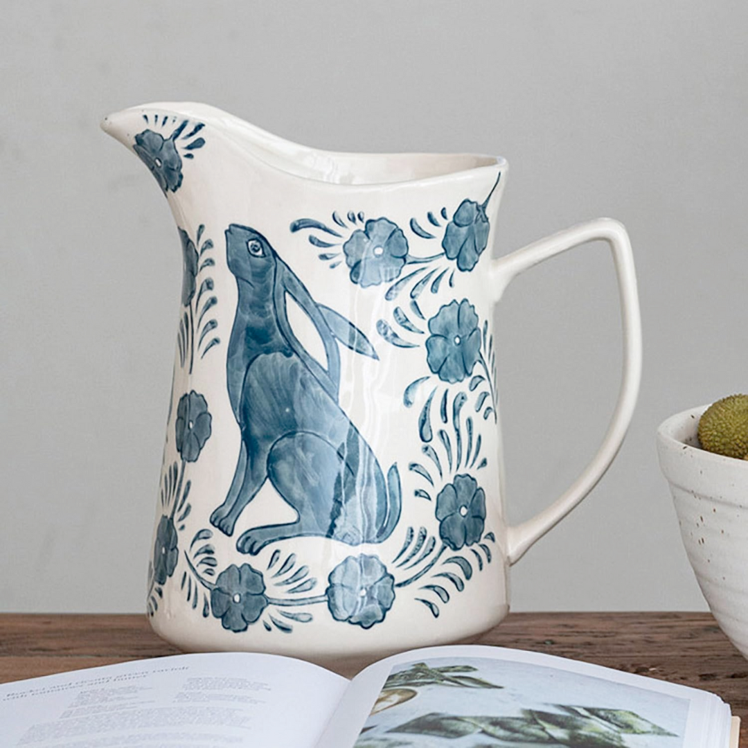 White stoneware pitcher with blue floral and rabbit design on a wooden surface.