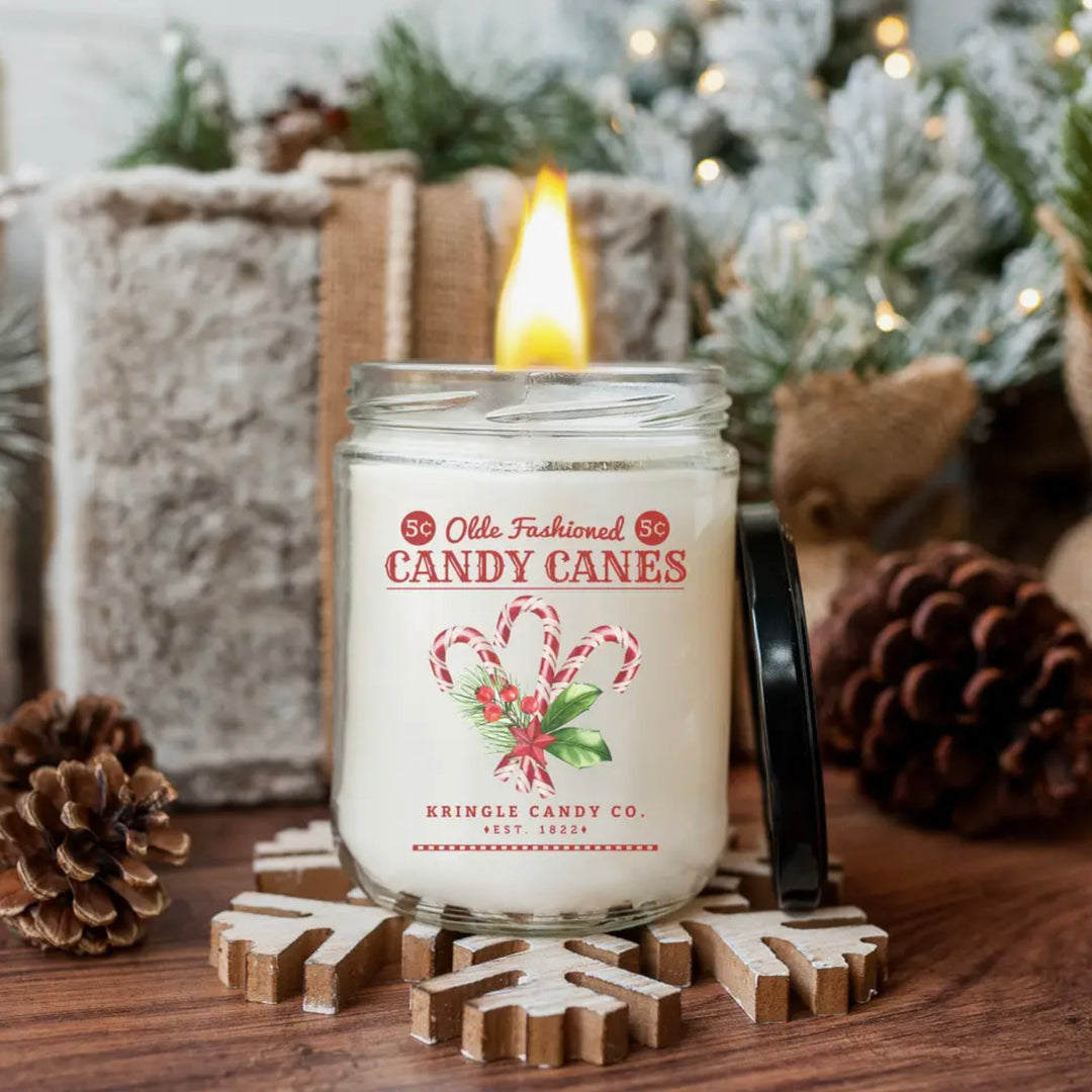 A blueberry candy cane scented candle with a label that reads 'Old Fashioned Candy Canes' placed in a glass jar with a black lid, surrounded by pine cones and decorative items.
