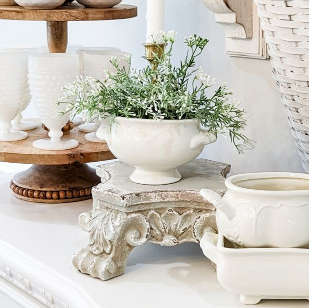 A white ceramic planter bowl filled with green and white baby's breath flowers, displayed on a distressed resin pedestal riser in a styled home decor setting.
